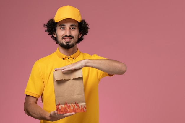 Front view male courier in yellow uniform holding paper food package and smiling on light-pink wall