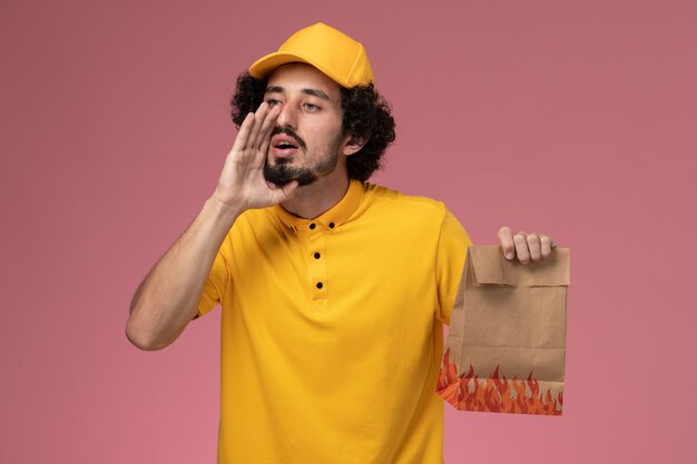 Front view male courier in yellow uniform holding paper food package whispering on light-pink wall