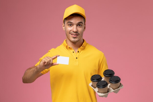 Front view male courier in yellow uniform holding plastic card and delivery coffee cups on the pink background