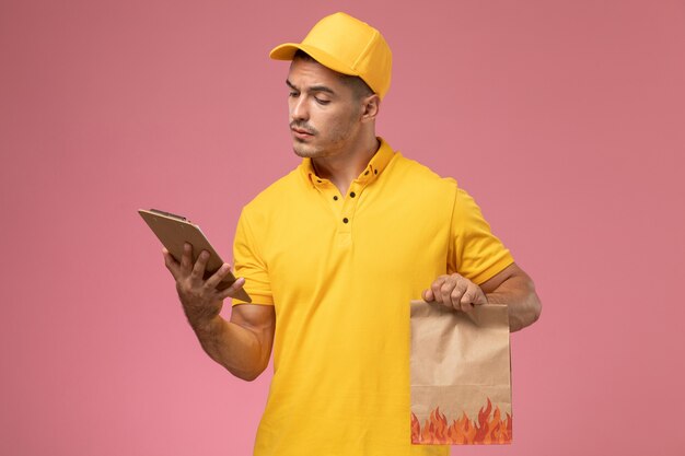 Front view male courier in yellow uniform reading notepad and holding food package on the pink desk 