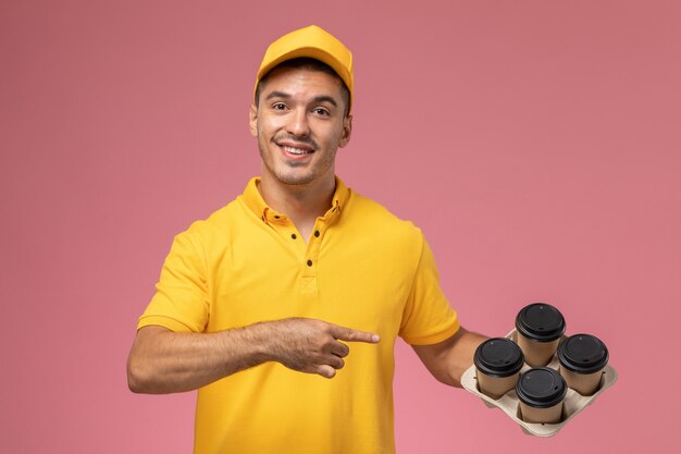 Front view male courier in yellow uniform smiling and holding delivery coffee cups on pink