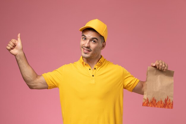 Front view male courier in yellow uniform smiling and holding food package on the pink background 