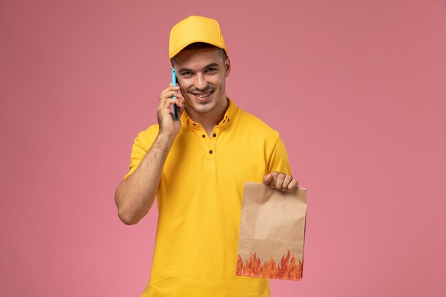 Вид спереди мужской курьер в желтой форме улыбается, разговаривает по телефону, держа пакет с едой на розовом