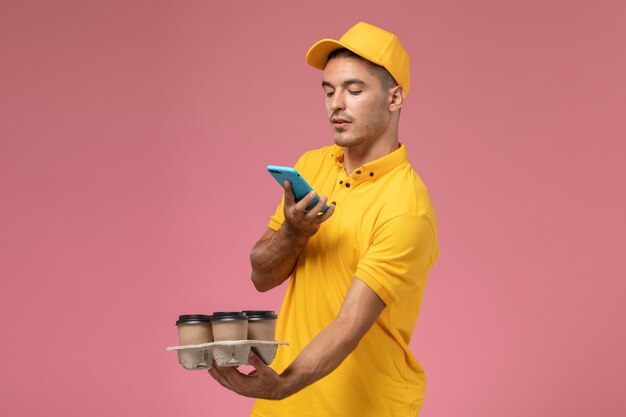 Front view male courier in yellow uniform taking a photo of delivery coffee cups on pink desk  