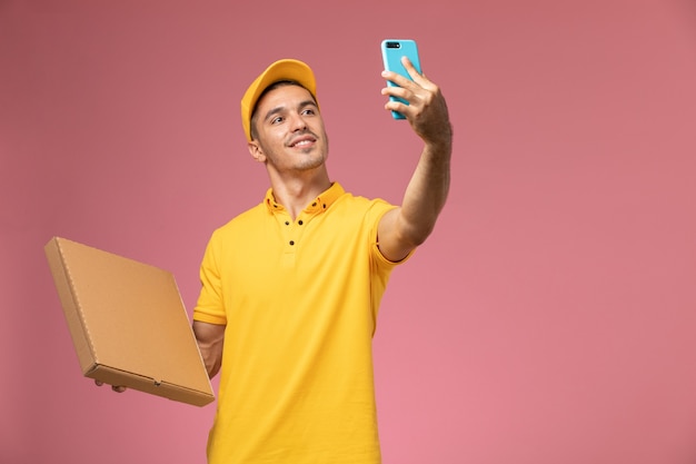 Курьер-мужчина в желтой форме, вид спереди, фотографирует с коробкой для доставки еды на розовом столе