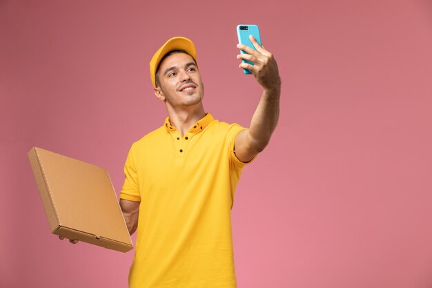 Front view male courier in yellow uniform taking a photo with food delivery box on pink desk 