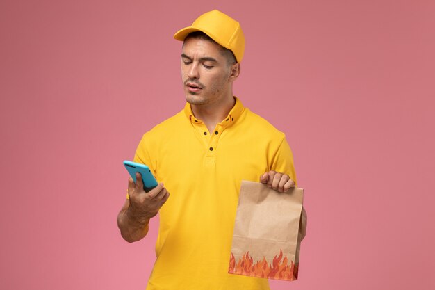 Курьер-мужчина в желтой форме, вид спереди, используя свой телефон, держа пакет с едой на розовом фоне