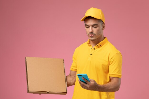 Курьер-мужчина в желтой форме, вид спереди, разговаривает по телефону и держит коробку для доставки еды на розовом столе