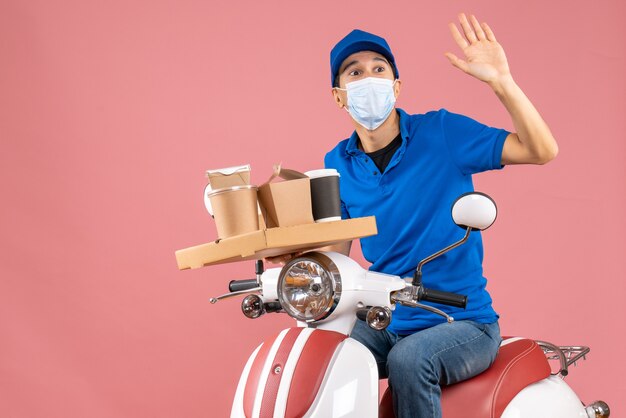 Front view of male delivery person in mask wearing hat sitting on scooter delivering orders on pastel peach background