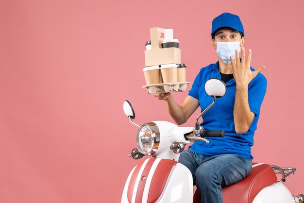 Front view of male delivery person in mask wearing hat sitting on scooter delivering orders showing ten on peach background