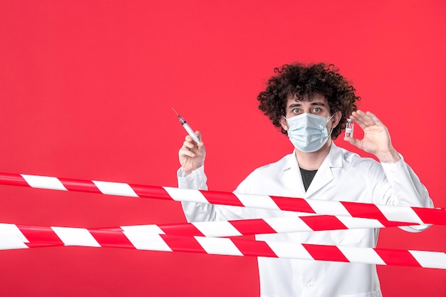 Бесплатное фото Врач-мужчина в медицинской форме, вид спереди, держит фляжку и инъекцию на красном фоне, карантинная полоса covid-предупреждение цвета больницы об опасности для здоровья