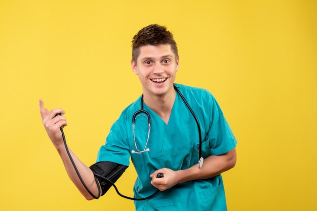 Front view of male doctor in medical suit checking his pressure on a yellow wall