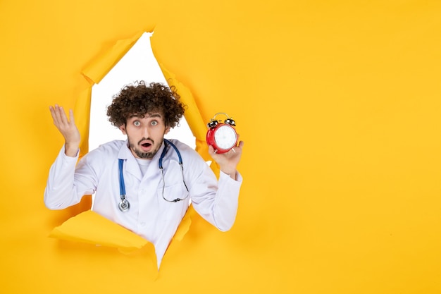 Front view male doctor in medical suit holding clocks on a yellow health color hospital shopping medicine time medic