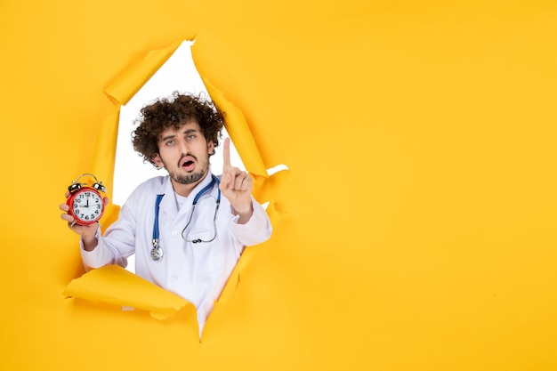 Вид спереди мужчина-врач в медицинском костюме, держащий часы на желтой больнице, шоппинг, медицина, цвет, время, здоровье