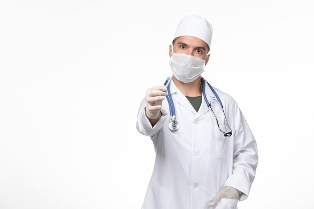 医療スーツを着た正面図の男性医師と、ライトホワイトウォールウイルス感染症パンデミックのコロナウイルス保持装置に対するマスク
