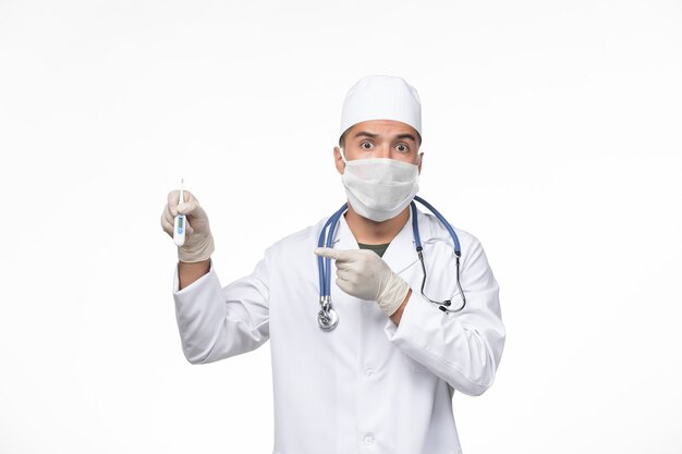 Front view male doctor in medical suit and mask against coronavirus holding device on the white wall virus covid- illness disease pandemic