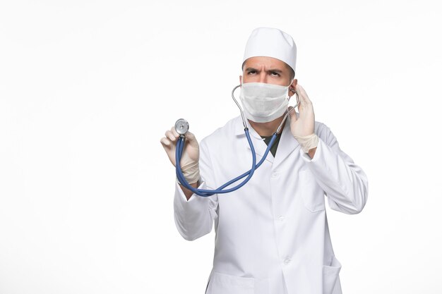 Front view male doctor in medical suit and mask against coronavirus using a stethoscope on the white wall covid- illness disease pandemic