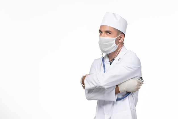 Front view male doctor in medical suit and mask against coronavirus using a stethoscope on the white wall covid- virus illness disease pandemic