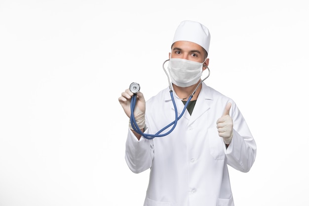 Front view male doctor in medical suit and mask against coronavirus using a stethoscope on white wall covid- virus illness disease pandemic