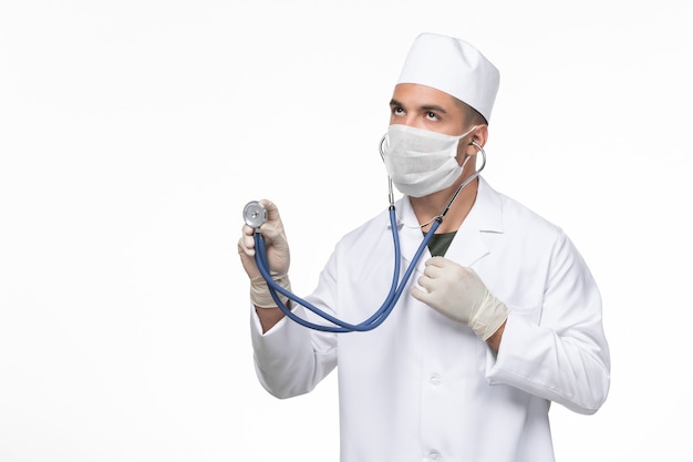 Front view male doctor in medical suit and mask against coronavirus using stethoscope on white wall covid- virus illness disease pandemic