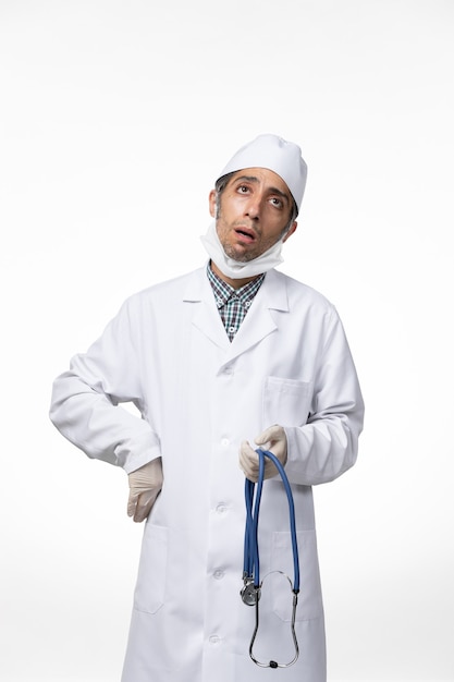 Вид спереди мужчина-врач в медицинском костюме и маске из-за коронавируса, держащего стетоскоп на белом столе