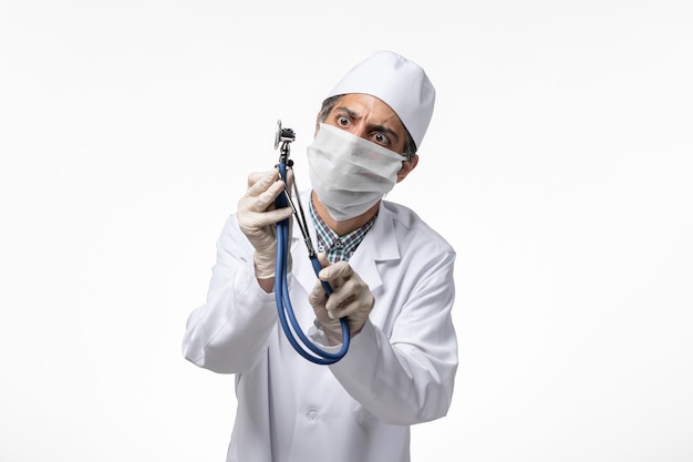 Free photo front view male doctor in medical suit and mask due to coronavirus holding stethoscope on white surface