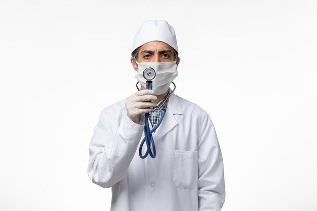 白い表面に聴診器を保持しているコロナウイルスによる医療スーツとマスクの正面図男性医師