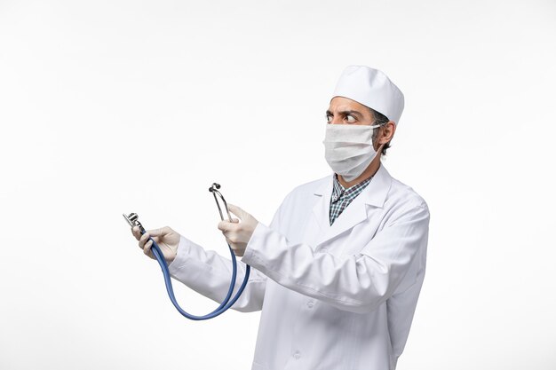 Front view male doctor in medical suit and mask due to coronavirus using stethoscope on light white surface