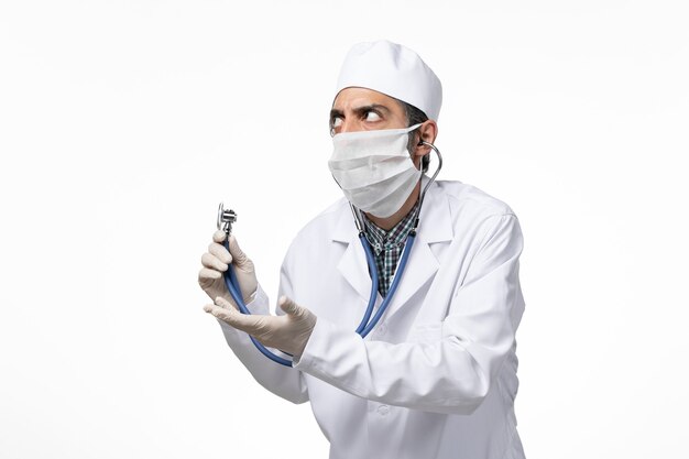 Front view male doctor in medical suit and mask due to coronavirus using stethoscope on light white surface