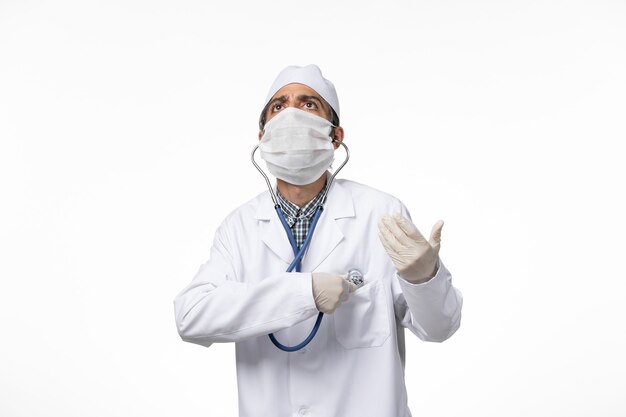 Front view male doctor in medical suit and mask due to coronavirus using a stethoscope on white desk