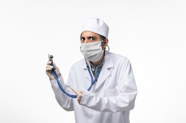 Front view male doctor in medical suit and mask due to coronavirus using a stethoscope on white desk