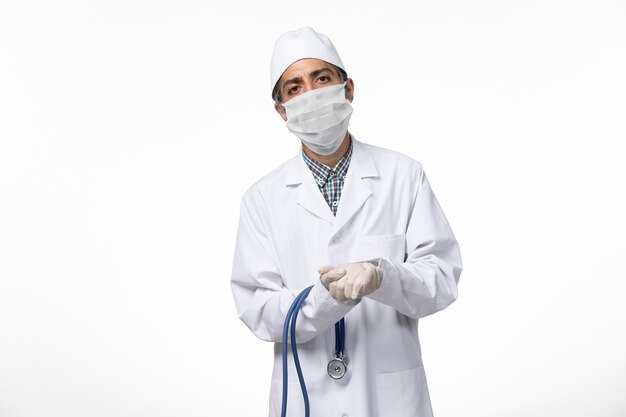 Вид спереди мужчина-врач в медицинском костюме и маске из-за коронавируса с использованием стетоскопа на белой поверхности