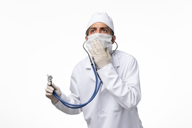 Front view male doctor in medical suit and mask due to coronavirus using a stethoscope on white surface