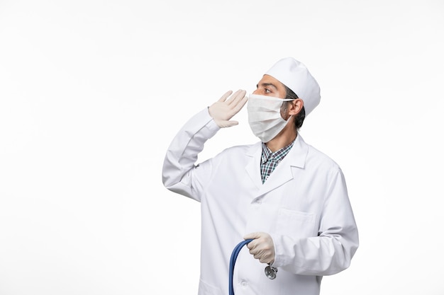 Front view male doctor in medical suit and mask due to coronavirus using stethoscope on a white surface