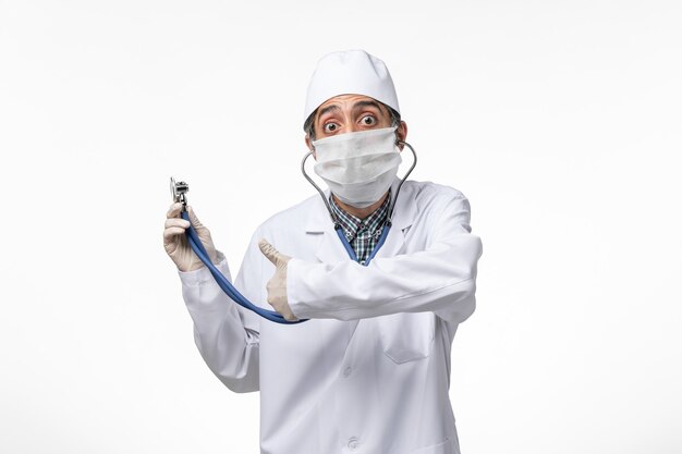 Front view male doctor in medical suit and mask due to coronavirus using stethoscope on a white surface