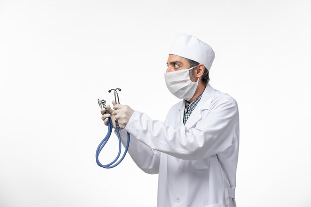 Front view male doctor in medical suit and mask due to coronavirus using stethoscope on a white surface