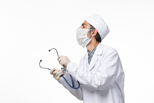 Front view male doctor in medical suit and mask due to coronavirus using stethoscope on white surface