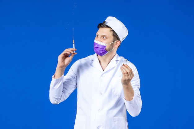 Front view of male doctor in medical suit and mask filling injection with vaccine on blue 