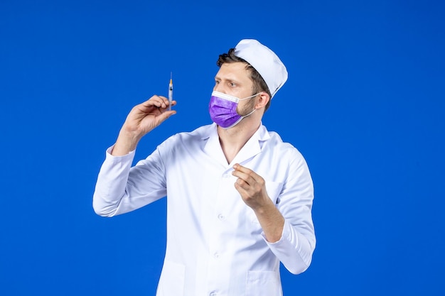 Front view of male doctor in medical suit and mask filling injection with vaccine on blue 