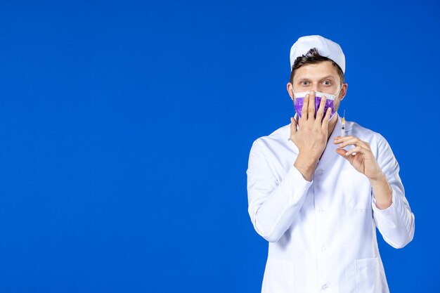 Front view of male doctor in medical suit and mask holding injection on blue 