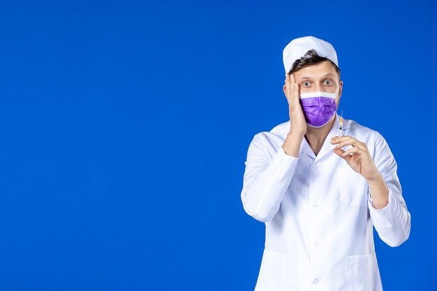 Front view of male doctor in medical suit and mask holding injection on blue 