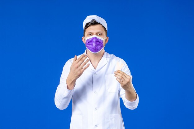 Front view of male doctor in medical suit and mask holding little medical patches on blue 