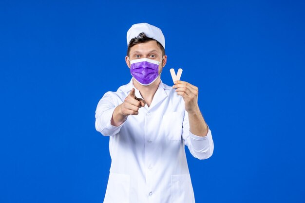 Front view of male doctor in medical suit and mask holding little medical patches and pointing at you on blue 