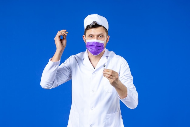 Front view of male doctor in medical suit and mask holding vaccine and injection on blue 