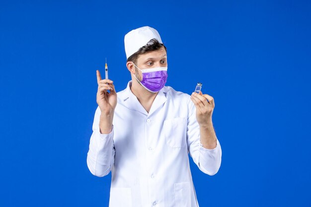 Front view of male doctor in medical suit and mask holding vaccine and injection on blue 