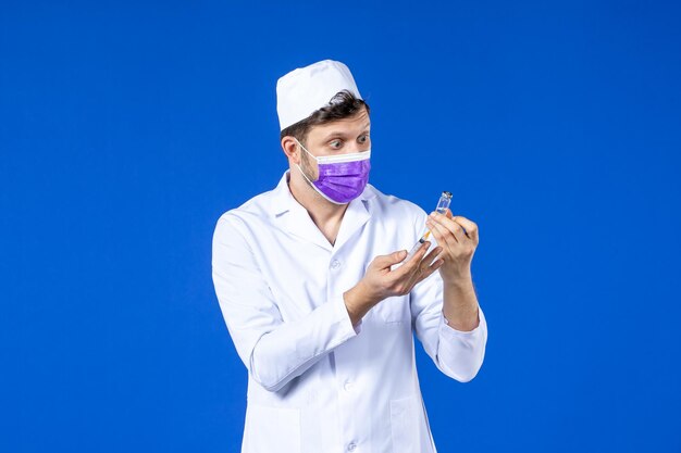 Front view of male doctor in medical suit and mask holding vaccine and injection on blue 