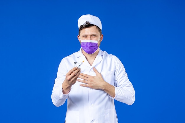 Front view of male doctor in medical suit and mask with injection and vaccine on blue 