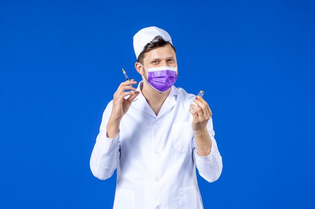 Front view of male doctor in medical suit and mask with injection and vaccine on blue 