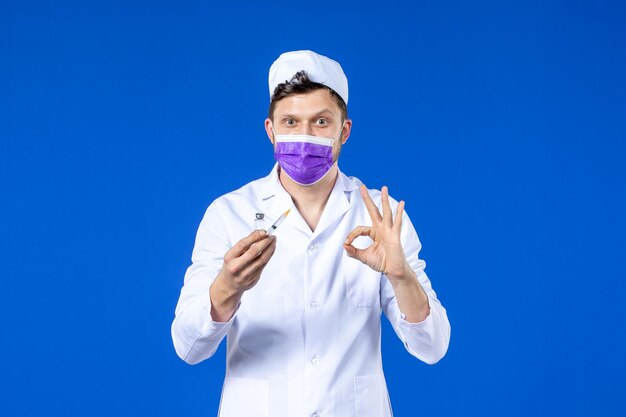 Front view of male doctor in medical suit and mask with injection and vaccine on blue 