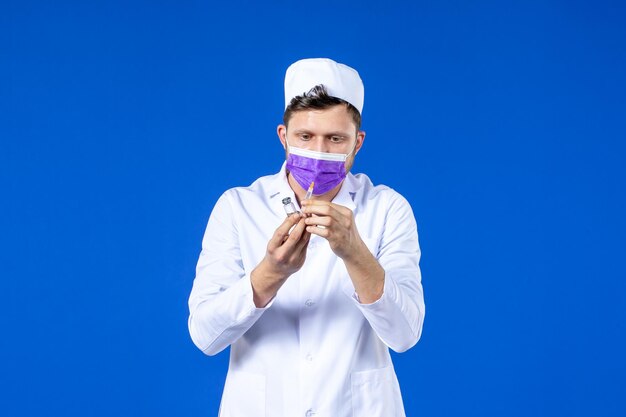 Front view of male doctor in medical suit and mask with injection and vaccine on blue 